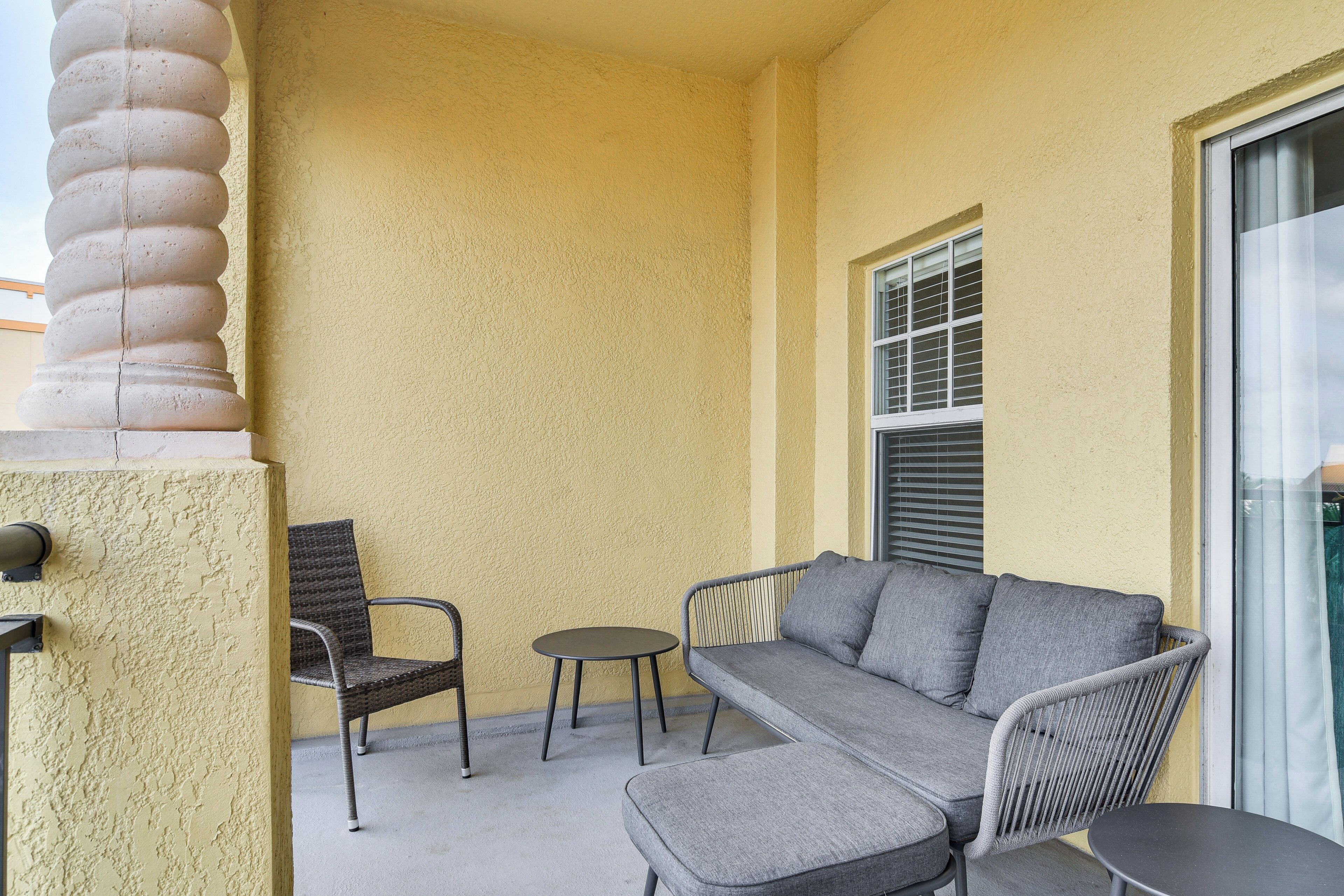 Ein Balkon mit einer grauen Couch, zwei Korbstühlen, einem kleinen runden Tisch und einem Fenster mit Jalousien.