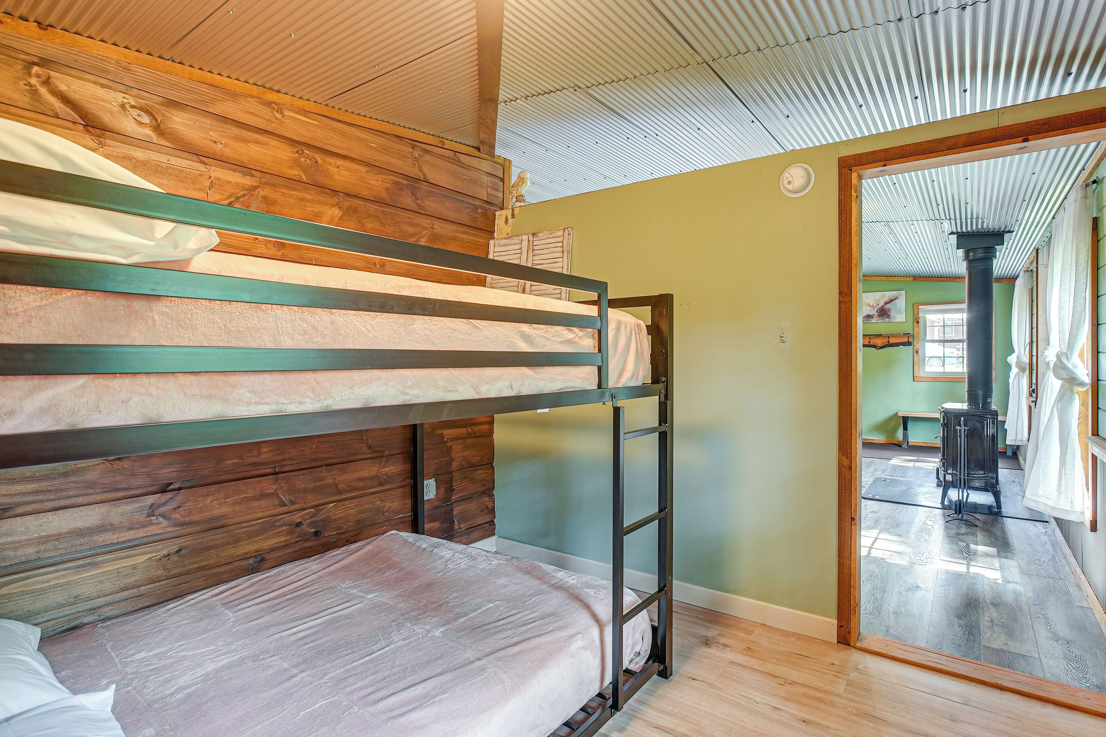 Un lit superposé avec une tête de lit en bois et une structure en métal, un plancher en bois et une vue sur une pièce où se trouve un poêle.