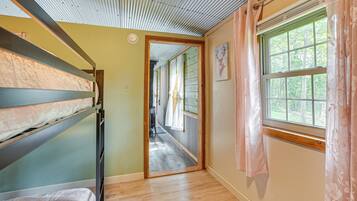 A room with bunk beds, a wooden floor, a window with curtains, and a door leading to another room.
