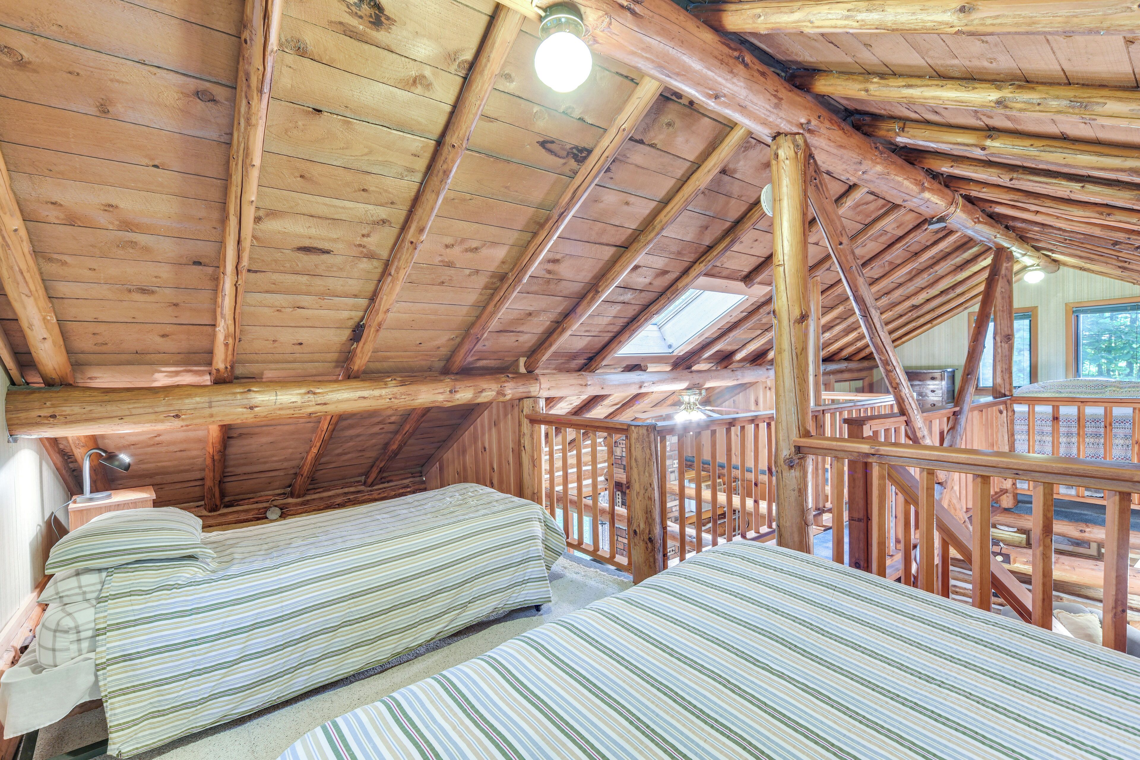A cabin with a sloped wooden ceiling, two beds with striped bedding, a wooden railing, and a window providing natural light.