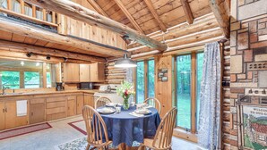 A wooden cabin interior with a dining table set for four, wooden chairs, a kitchen area, and large windows. - Authentic Log Cabin w/ Private Pond in Glidden (Glidden)