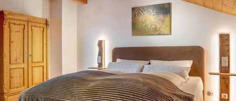 A bedroom with a wooden ceiling, a bed with a striped blanket, a wooden wardrobe, and a wall-mounted light fixture.
