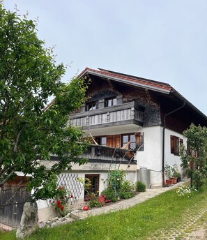 A two-story house with a balcony, a garden with flowers, and a tree in the foreground. - Cozy Apartment with Mountain Views and Box Spring Bed (Pfronten)