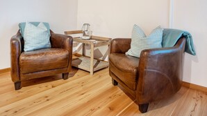 Deux fauteuils en cuir agrémentés de coussins à carreaux, une petite table en bois sur laquelle se trouve une carafe en verre, et un plancher en bois.