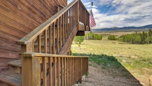 A wooden cabin with a wooden staircase leading to a porch, an American flag, and a view of a grassy field and mountains in the background. - Private 5 Acres w/ Mtn Views: Park County Cabin! (Jefferson)