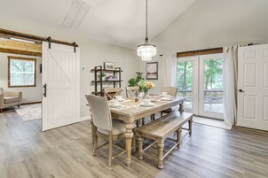 Comedor con mesa de madera para cuatro personas, una araña y vistas a través de amplios ventanales.