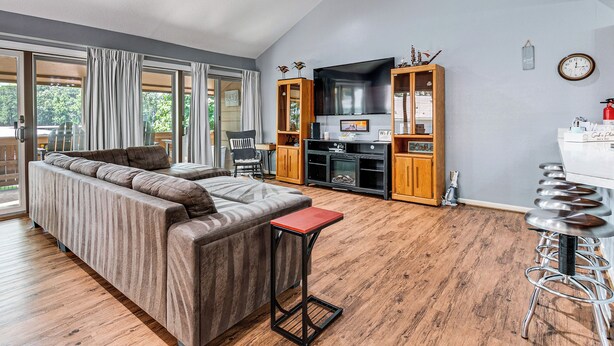 A modern living room with a sectional sofa, a small table, and a bar area with stools. There is a flat-screen TV mounted above the fireplace, wooden cabinets, and a large window with curtains.