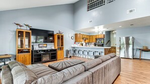 A spacious living area with a large gray sectional sofa, wooden cabinets, and a kitchen with a stainless steel refrigerator.