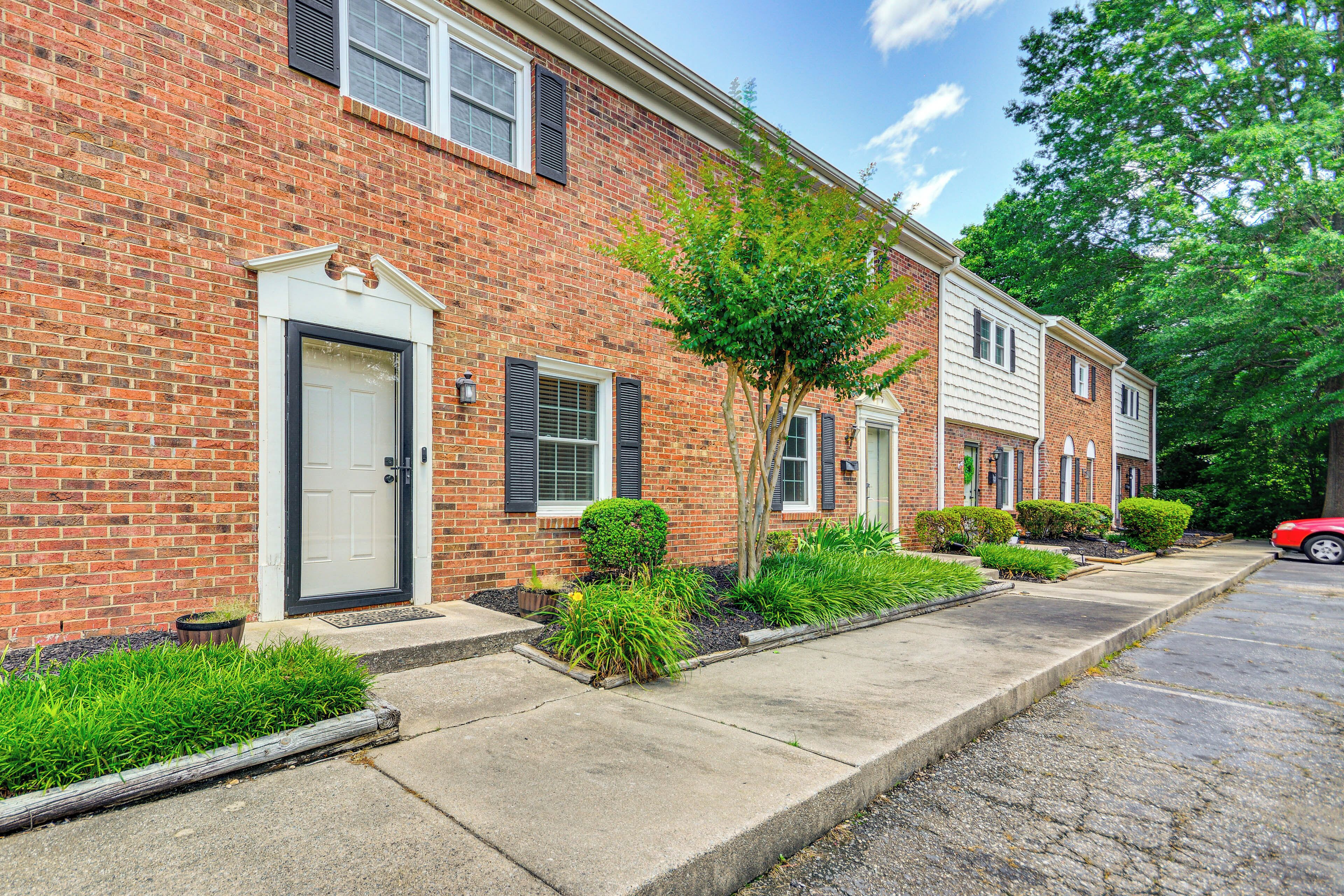 A row of brick townhouses with front doors, small front yards with plants, and a red car parked on the street.