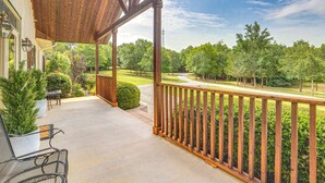 Une véranda couverte avec une balustrade en bois, des meubles d’extérieur et une vue sur un jardin et des arbres bien entretenus.