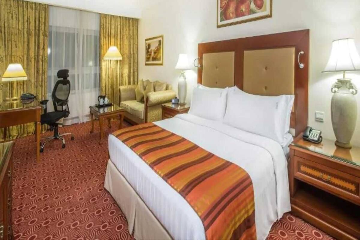 A hotel room with a large bed, two bedside tables, a desk, a chair, and a large window with curtains.