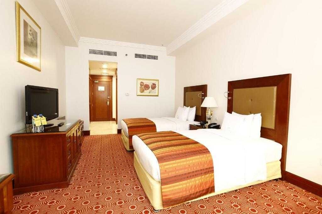 A hotel room with two beds, a TV, a desk, and a patterned carpet.