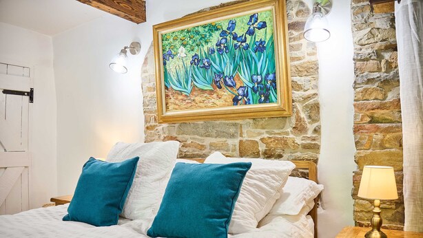 A bed with white bedding and two teal pillows, a painting of irises on the wall, a stone wall, and a nightstand with a lamp. - Host Stay Dipple Farm Cottage (Bideford)