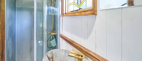 A bathroom with a glass shower enclosure, a metal bucket, and a wooden-framed window.