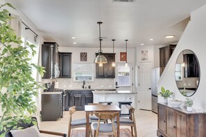 A modern kitchen with dark cabinets, a central island, pendant lighting, and a dining area with wooden chairs and a table. - Luxury 4BR 2 5BA Home EV Charger Pool Access (Buda)