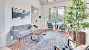 A spacious living room with a grey sofa, a wooden coffee table, and a patterned rug. There is a ceiling fan, a large window with curtains, and a potted plant. - Luxury 4BR 2 5BA Home EV Charger Pool Access (Buda)