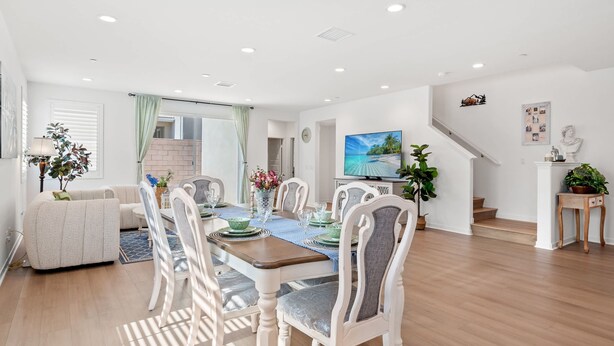 A spacious living area with a dining table set for six, surrounded by white upholstered chairs. A large flat-screen TV is mounted on the wall, and a staircase leads to an upper level. - Spacious 5BR Getaway w Game Room Modern Comfort (Fontana)