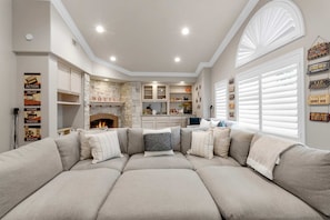A living room with a sectional sofa, a fireplace, built-in shelving, and a kitchen area in the background.