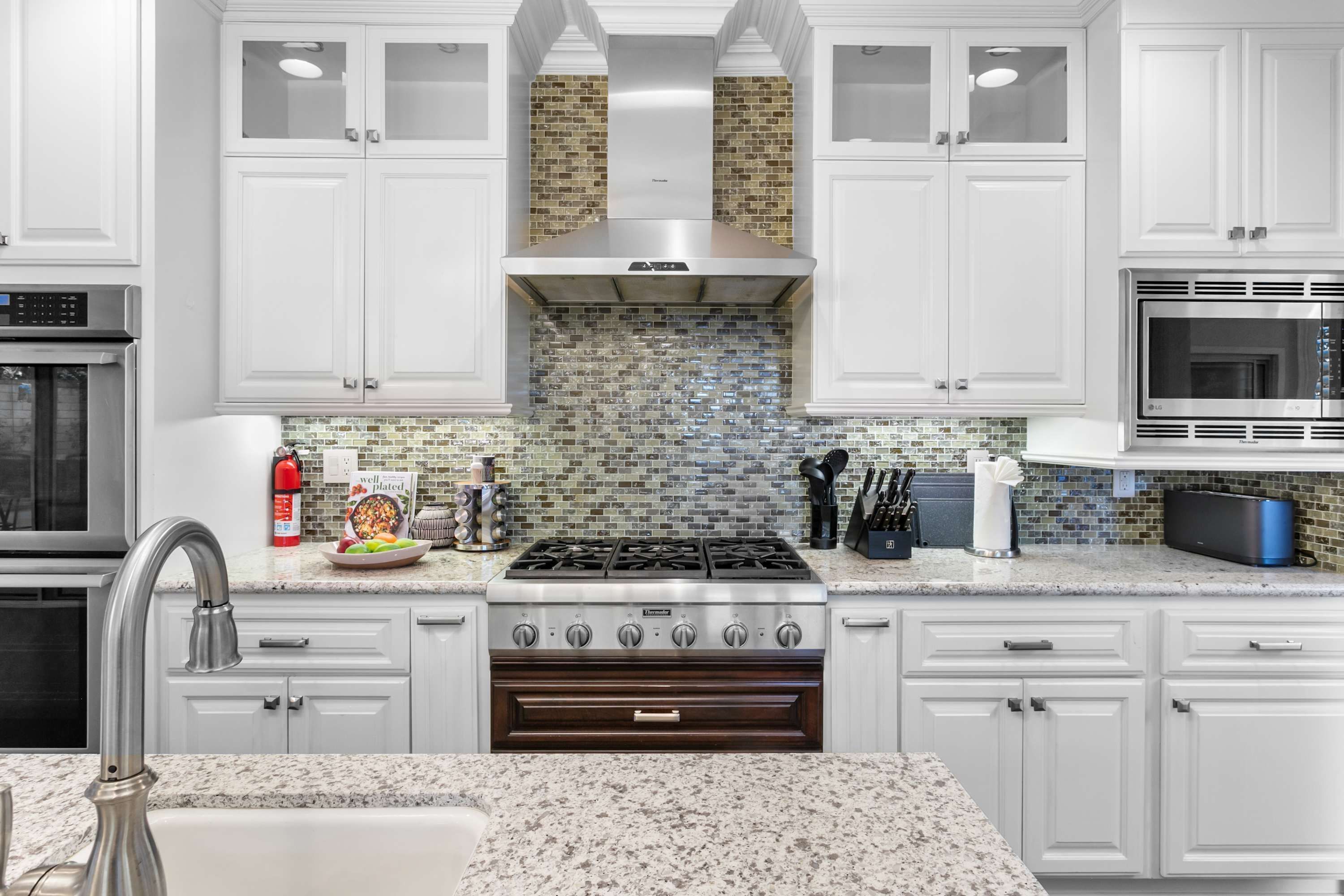 Una cocina moderna con armarios blancos, estufa de acero inoxidable, horno integrado y salpicadero con mosaicos.