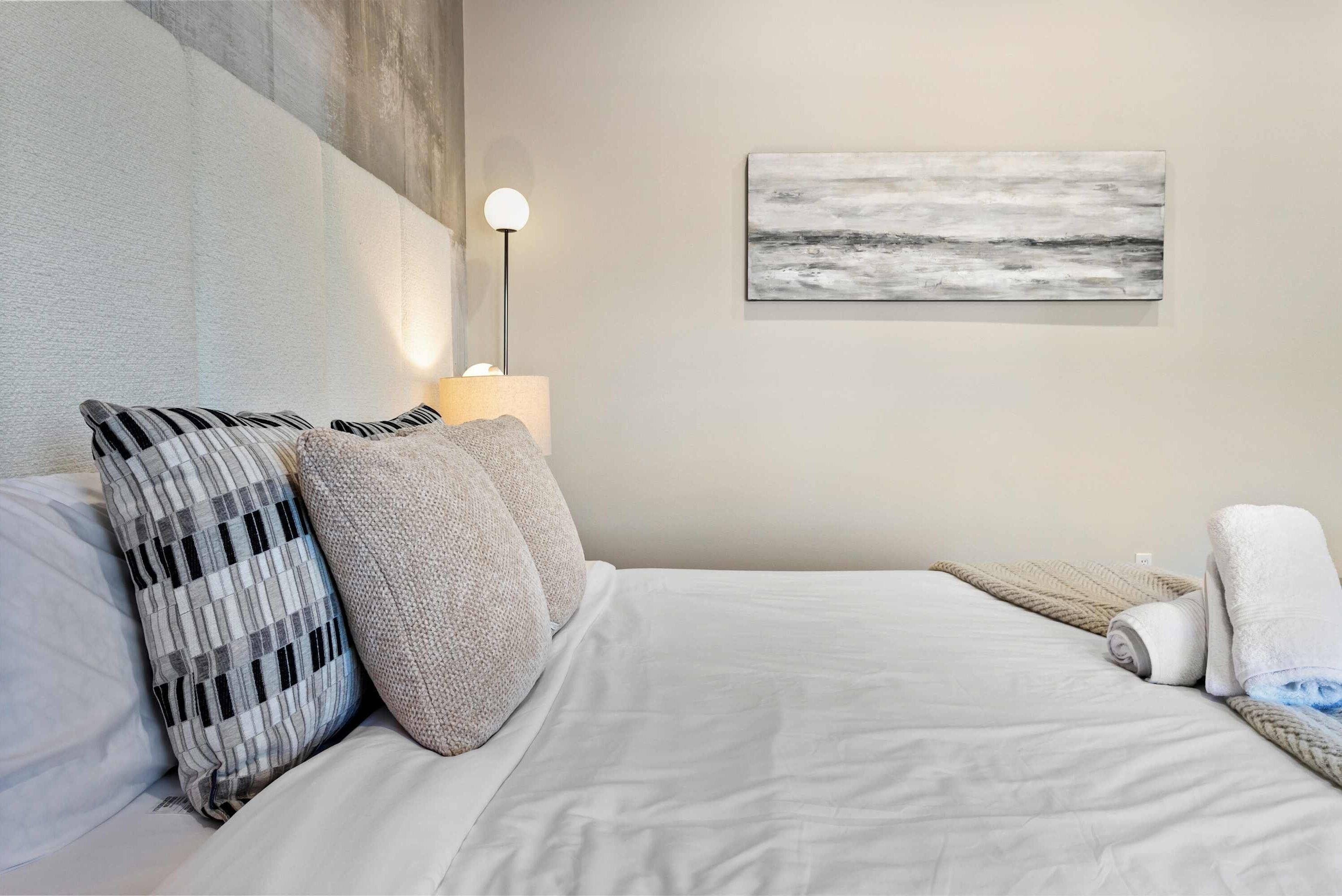 A neatly made bed with white linens, two pillows, a lamp, and a painting on the wall.