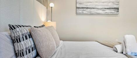 A neatly made bed with white linens, two pillows, a lamp, and a painting on the wall.
