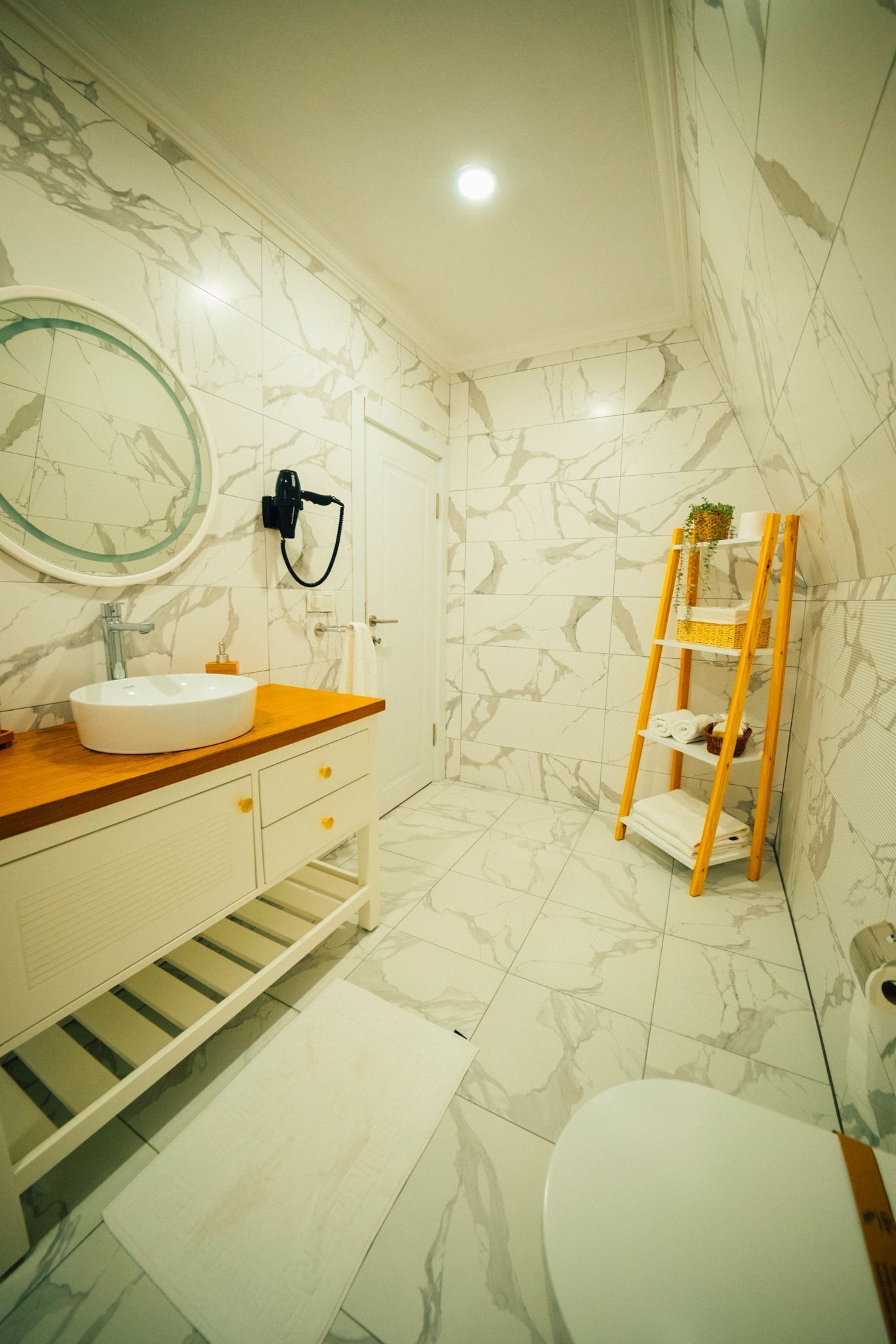 Une salle de bain avec un lavabo blanc, un meuble-lavabo en bois, des carreaux de marbre, une étagère en forme d’échelle en bois et des toilettes.
