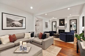 A modern living room with a grey sectional sofa, a blue accent chair, a fireplace, and a large framed landscape painting. - Renovated Victorian Home | Forsyth Park (Savannah)