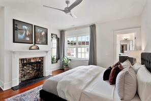 A bedroom with a fireplace, a bed with multiple pillows, a ceiling fan, and a large window with curtains. - Renovated Victorian Home | Forsyth Park (Savannah)