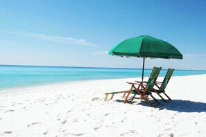 A beach with white sand and turquoise water. There is a green umbrella and two wooden beach chairs.
