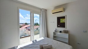 Una habitación de hotel con cama, ventana con cortinas, aire acondicionado de pared, un cuadro enmarco y una pequeña estantería con objetos decorativos.