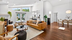A spacious living area with a large sofa, coffee table, and dining table. There is a kitchen area in the background, a ceiling fan, and a view of the outdoors through large windows.