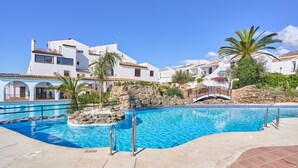 Piscina com uma ilha de pedra, edifícios brancos com varandas e um céu azul límpido.