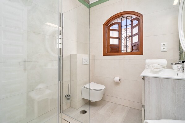 A modern bathroom with a glass shower enclosure, a toilet, a sink with a mirror, and a window with a wooden frame.