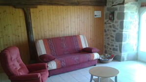 A room with a wooden wall, a stone wall, a red armchair, a patterned sofa, and a round table. - Holiday Home 'Gîte Le Rosier' with Wi-Fi (Saint-Mamet-la-Salvetat)