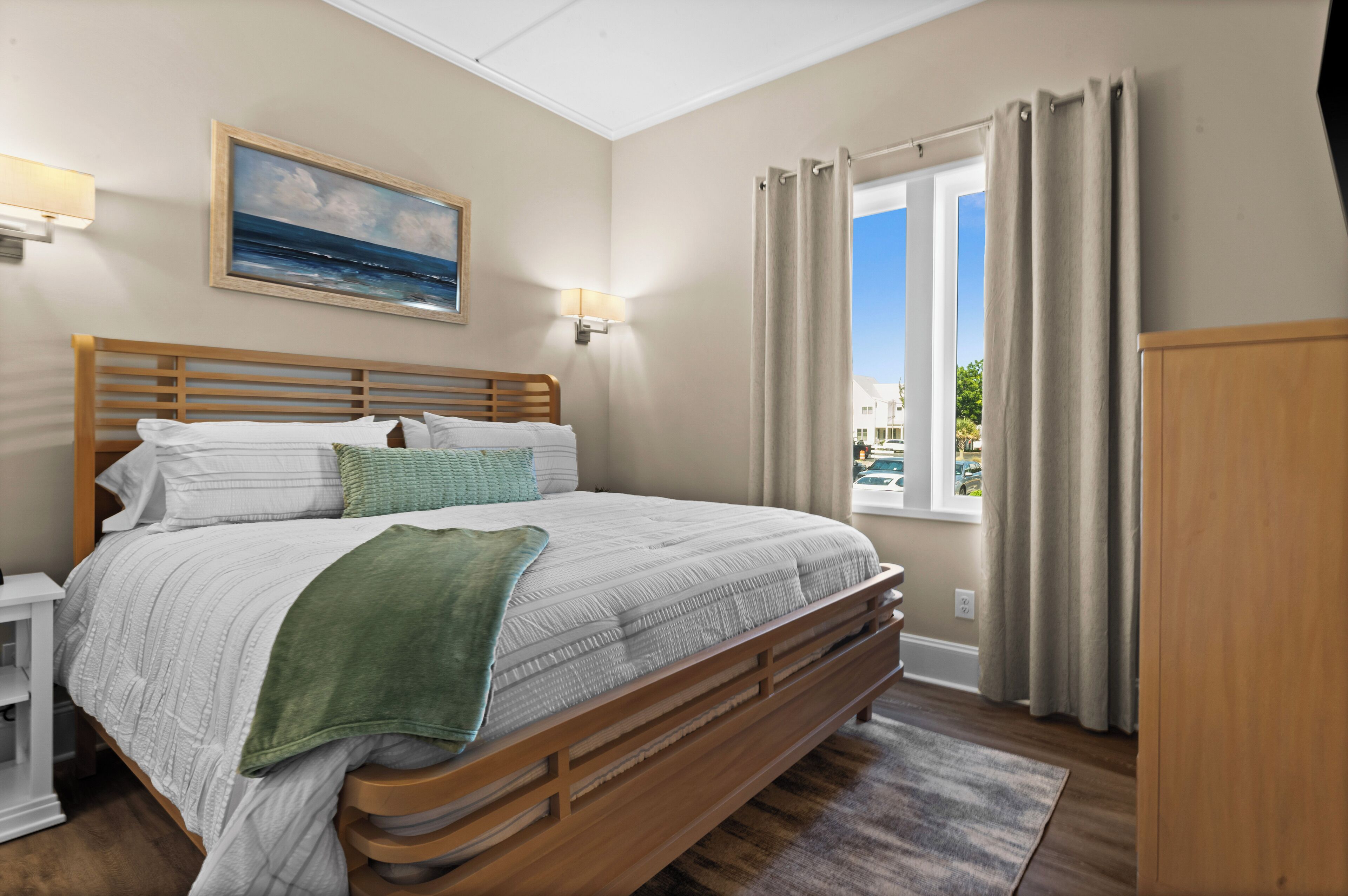 A neatly arranged bedroom with a wooden bed, a painting on the wall, and a window with curtains.