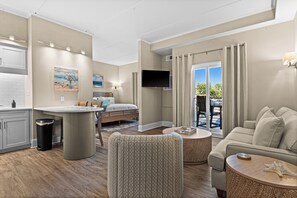 Una habitación de hotel moderna con kitchenette, zona de comedor, sala de estar con sofá y sillones, televisor de pantalla plana y un ventanal con cortinas.