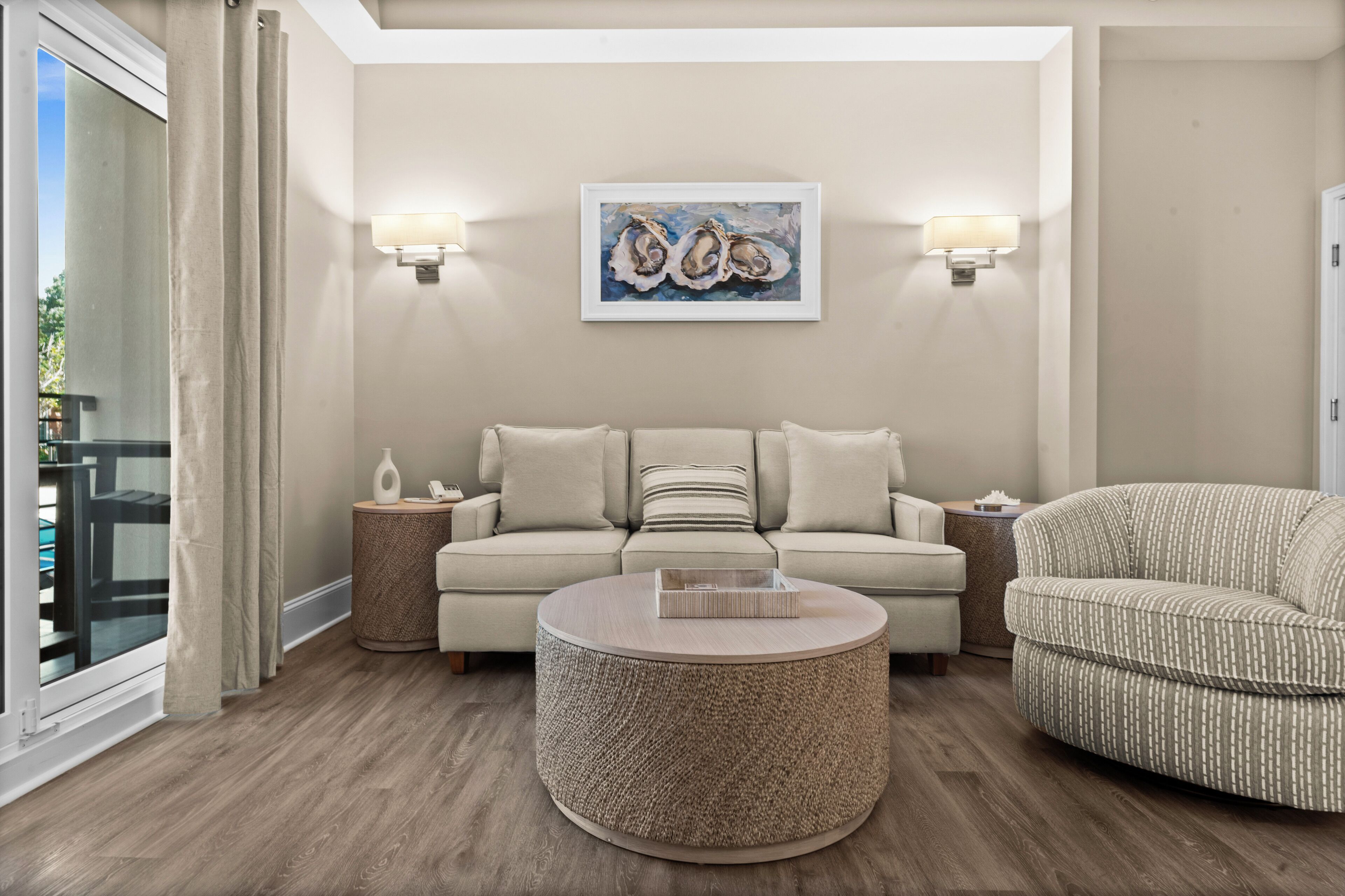 A living room with a beige sofa, a round coffee table, and a patterned armchair. There are two wall-mounted lamps and a framed artwork above the sofa.