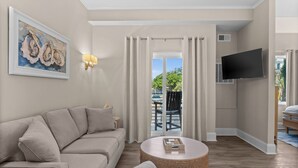 Un salon moderne avec un canapé beige, une table basse ronde, un téléviseur à écran plat fixé au mur et une grande fenêtre avec des rideaux.