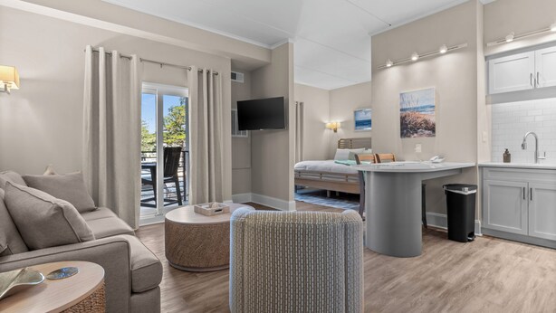Une chambre d’hôtel moderne équipée d’un canapé, d’un pouf, d’un coin repas, d’une kitchenette et d’un téléviseur fixé au mur.