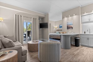 Une chambre d’hôtel moderne équipée d’un canapé, d’un pouf, d’un coin repas, d’une kitchenette et d’un téléviseur fixé au mur.