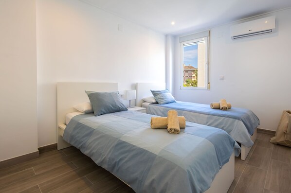 Habitación de hotel con dos camas, cada una con colcha azul y cabecero blanco. Se ve una ventana que deja entrar luz natural y un equipo de aire acondicionado en la pared.