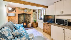 A cozy living room with a fireplace, a sofa with floral cushions, a television, and a kitchen area with cabinets and a microwave.
