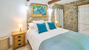 A bedroom with a stone wall, a bed with white bedding and blue pillows, a wooden nightstand with a lamp, and a wicker basket.
