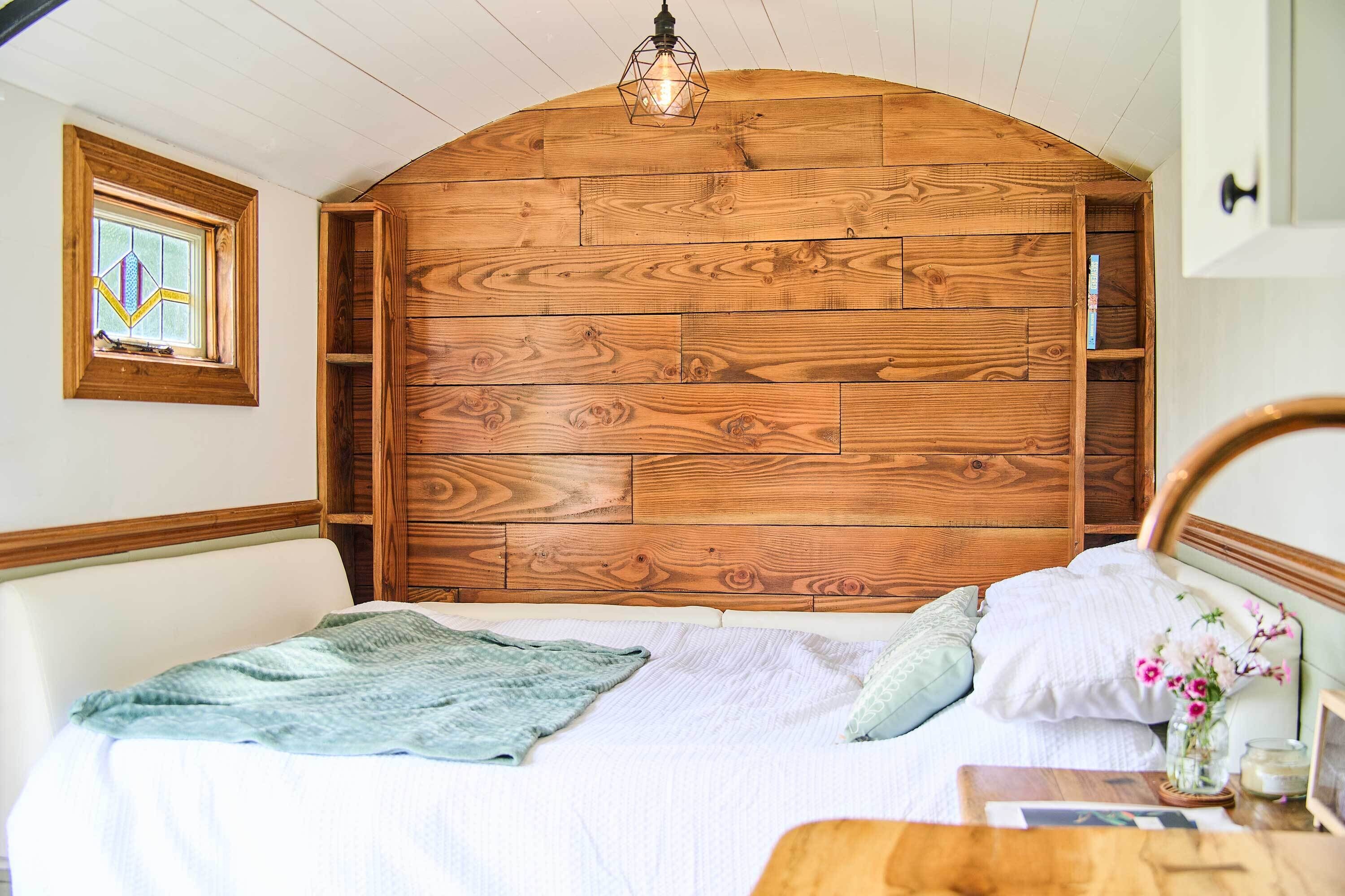Une chambre à coucher avec une tête de lit en bois, une fenêtre ornée de vitraux et une table de chevet sur laquelle est posé un vase de fleurs.