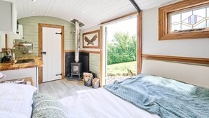 A cozy bedroom with a bed, a wooden desk, a fireplace, and a large window with a view of the outdoors. - Host & Stay | Dipple Farm Shepherds Hut (Woolfardisworthy)