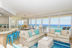 A spacious living area with a large window overlooking the ocean, a kitchen with a bar area, and a seating arrangement with blue stools.