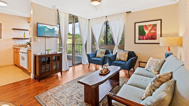 Ein geräumiges Wohnzimmer mit einem großen Sofa, einem Couchtisch und einem Fernseher. Es gibt eine Küchenzeile mit Herd und Schränken sowie einen Balkon mit Vorhängen.
