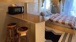 Interior acogedor de una cabaña con techo de madera, una cama con una colcha estampada, una pequeña cocina con microondas y un taburete de bar.