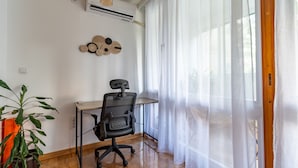 A room with a desk, chair, air conditioning unit, and a window with sheer curtains.
