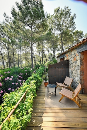 Une terrasse en bois avec deux fauteuils en osier et une table, entourée d’une végétation luxuriante et de plantes en fleurs.